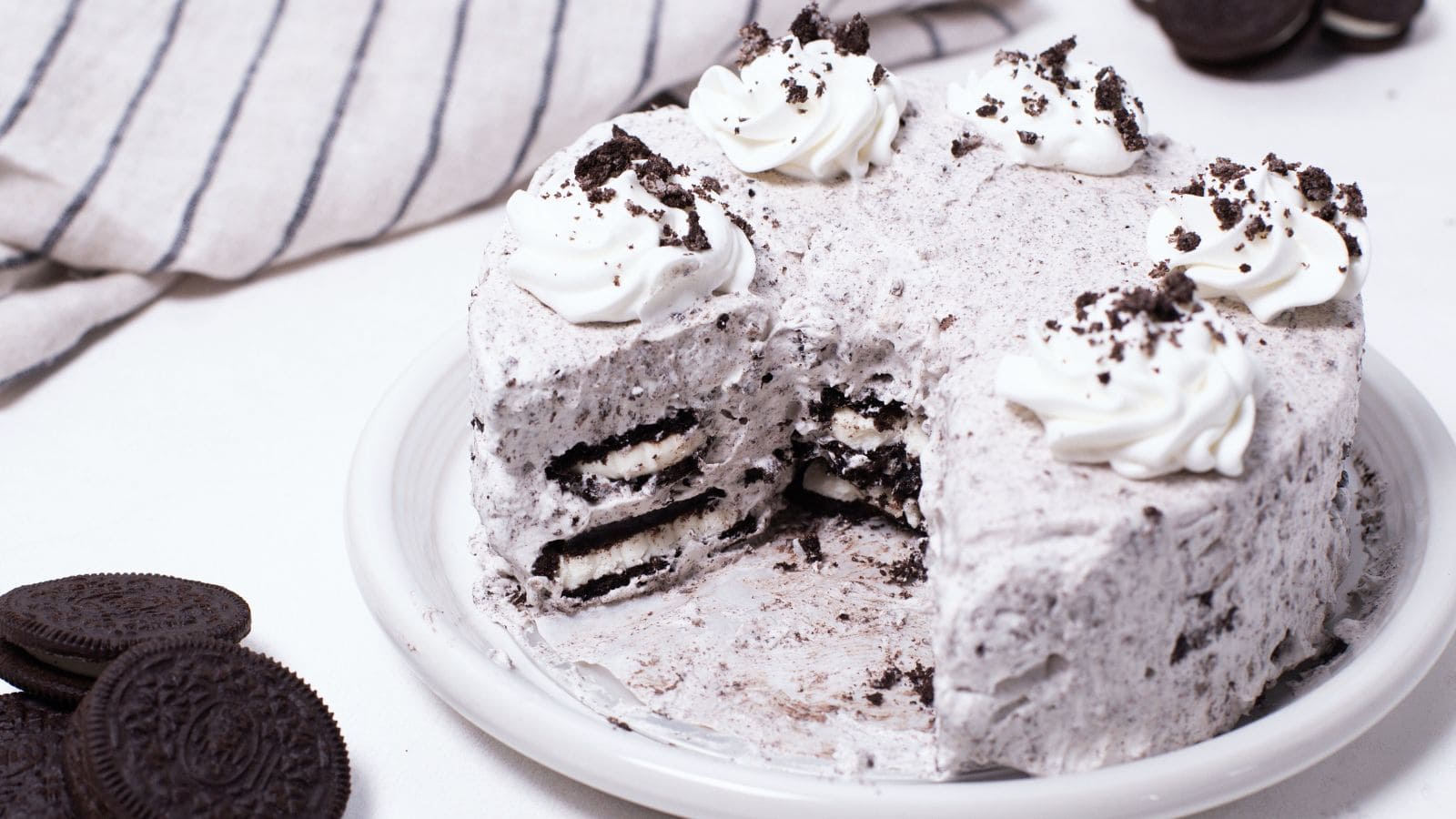 Cake with cream frosting topped with cookie crumbs and whole cookies inside, partially sliced, on a plate.
