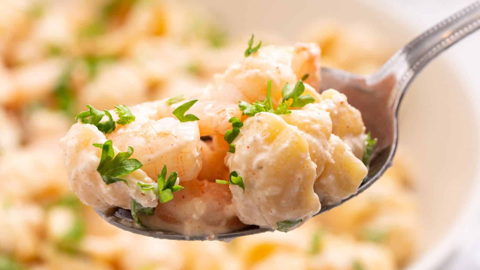 Close-up of a spoon holding seafood pasta salad garnished with parsley, with more salad in the background.