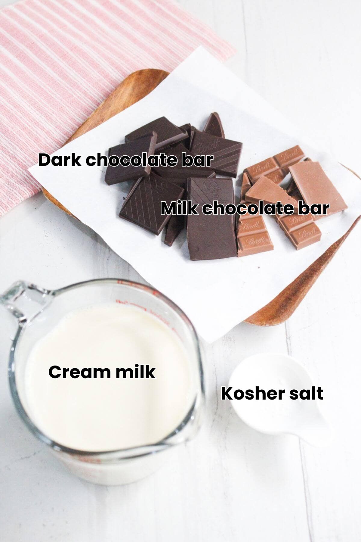 Chopped dark and milk chocolate on parchment paper with a glass measuring cup of cream and a small white spoon on a white surface.