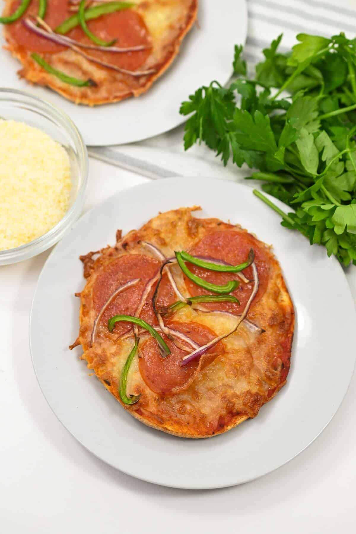 A plate with a mini pizza topped with pepperoni, green bell pepper, and red onion. Nearby is a bowl of grated cheese and fresh parsley.