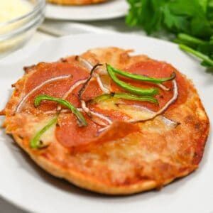 A small pizza topped with slices of pepperoni, green bell peppers, and onions on a white plate, with parsley in the background.