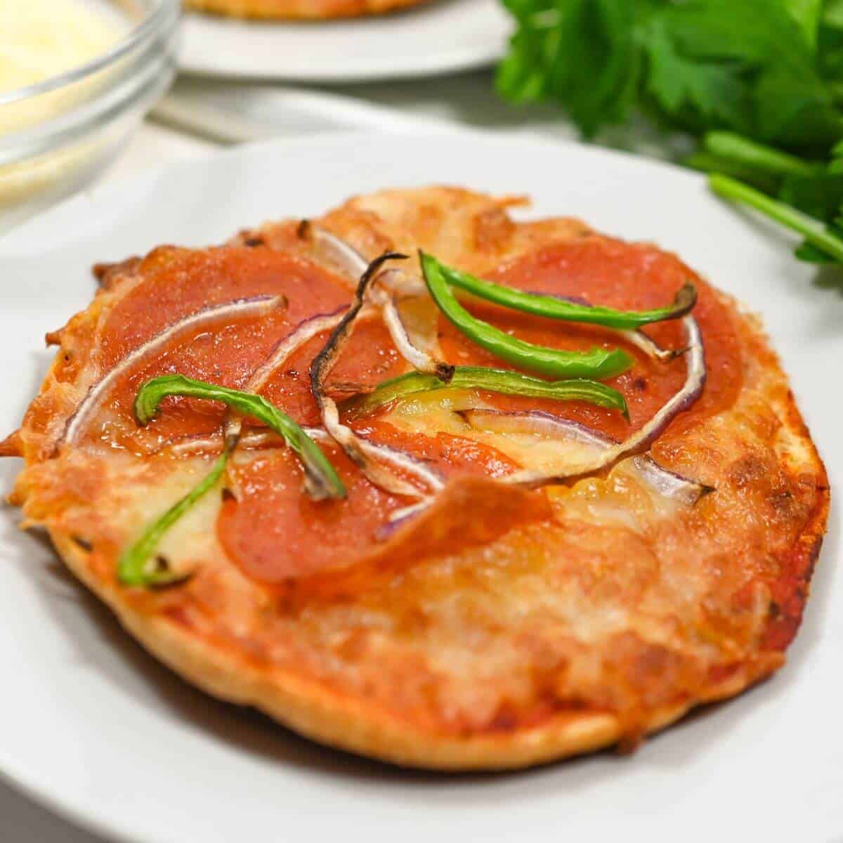 A small pizza topped with slices of pepperoni, green bell peppers, and onions on a white plate, with parsley in the background.