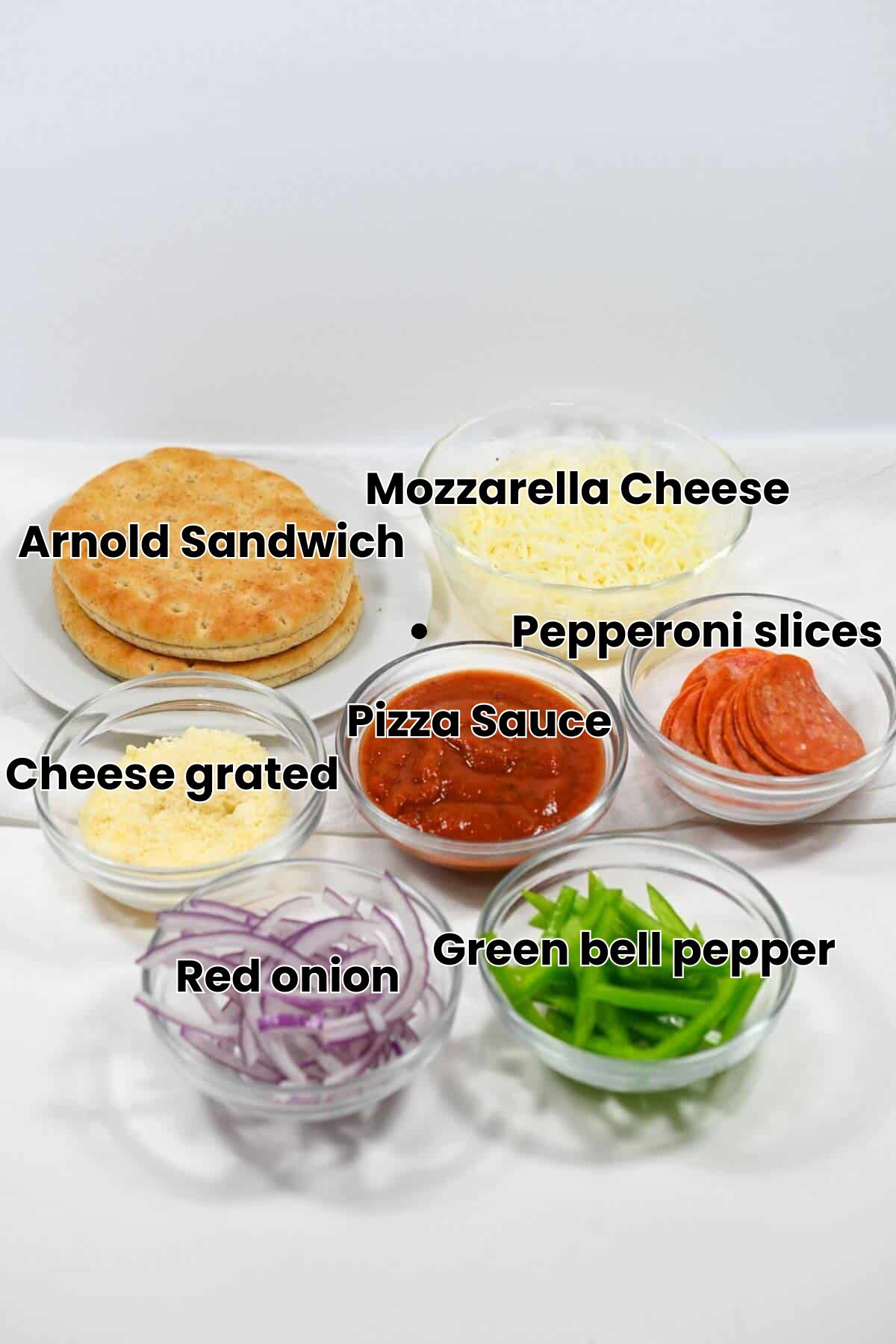 Various pizza ingredients in bowls: flatbread, shredded cheese, pepperoni slices, tomato sauce, grated parmesan, sliced red onions, and sliced green bell peppers.
