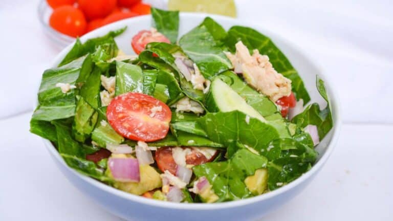 A bowl of fresh salad with leafy greens, cherry tomatoes, cucumber slices, red onion, and chunks of tuna. A lime wedge and cherry tomatoes are in the background.