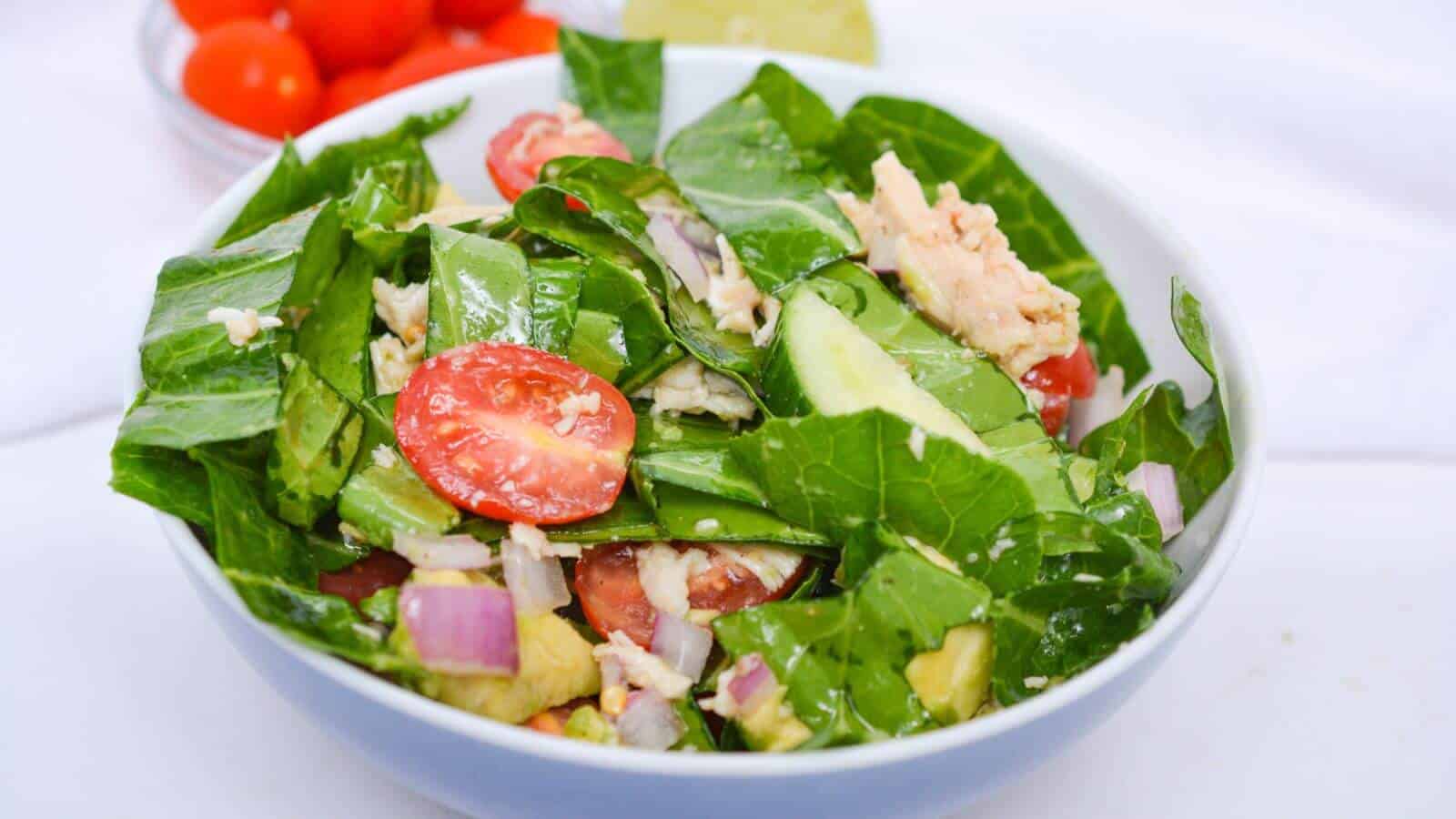 A bowl of fresh salad with leafy greens, cherry tomatoes, cucumber slices, red onion, and chunks of tuna. A lime wedge and cherry tomatoes are in the background.