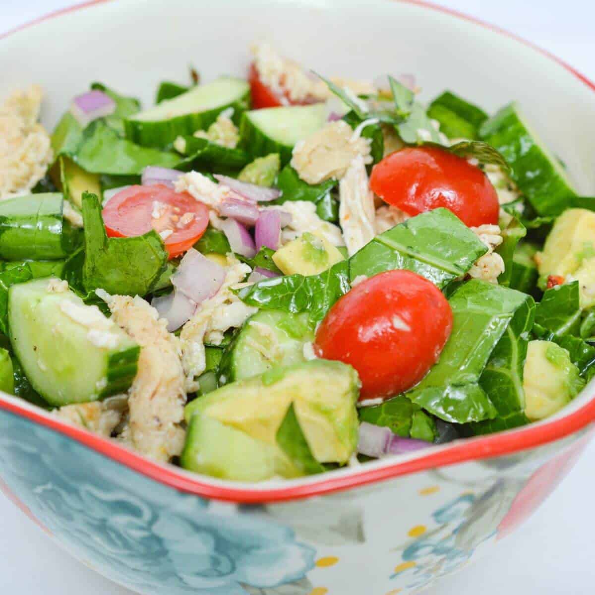 A colorful salad with chopped lettuce, cherry tomatoes, cucumber, avocado, red onion, and shredded chicken in a floral-patterned bowl.