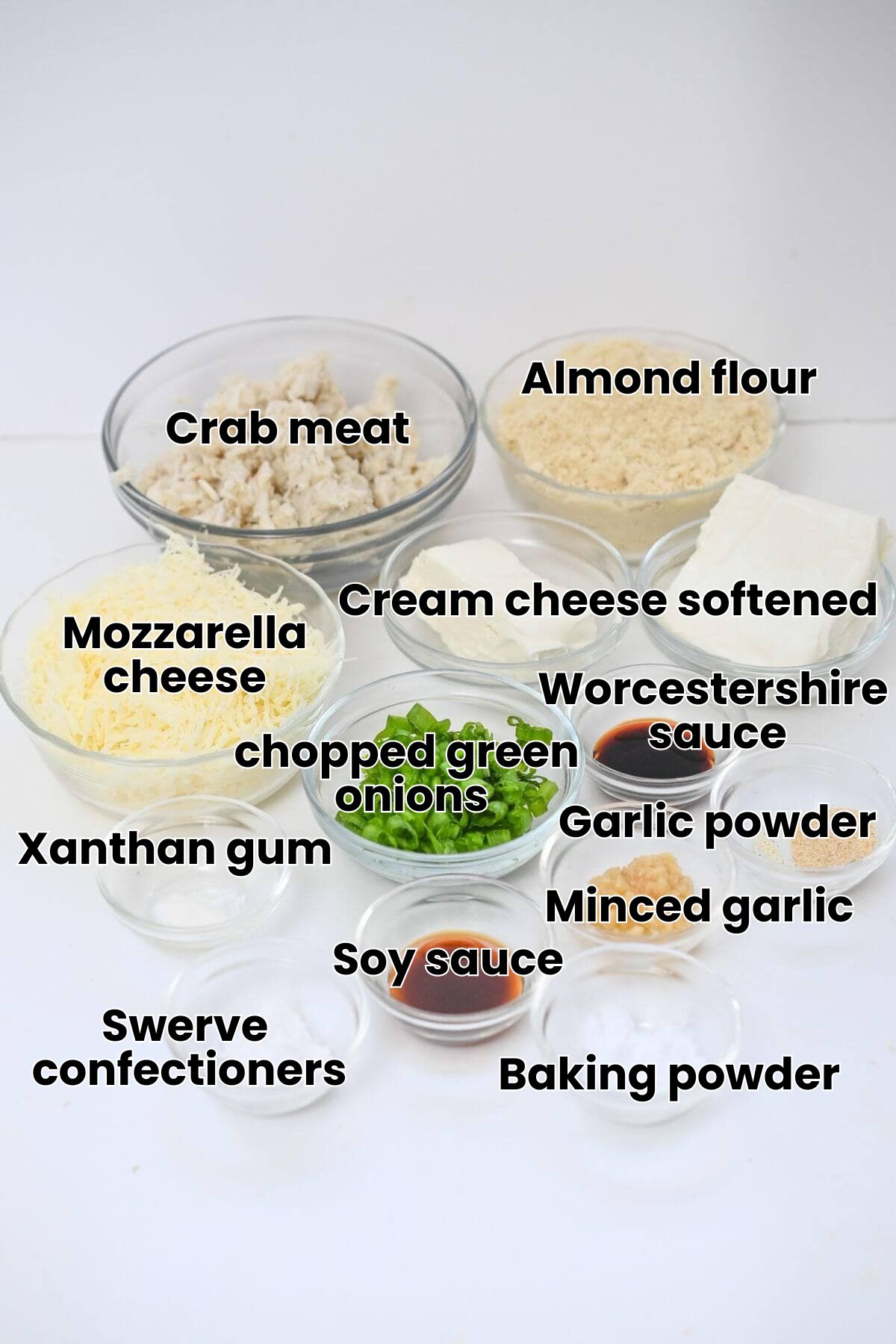Various ingredients in glass bowls including shredded cheese, chopped green peppers, chicken, bread crumbs, cream cheese, soy sauce, garlic, and seasonings arranged on a white surface.