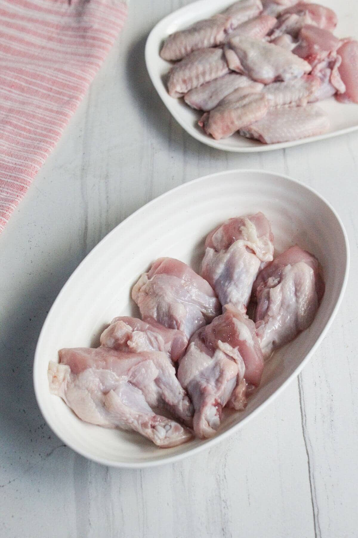 Two white oval plates with raw chicken wings are placed on a light-colored surface, with a striped cloth partially visible in the upper left corner.