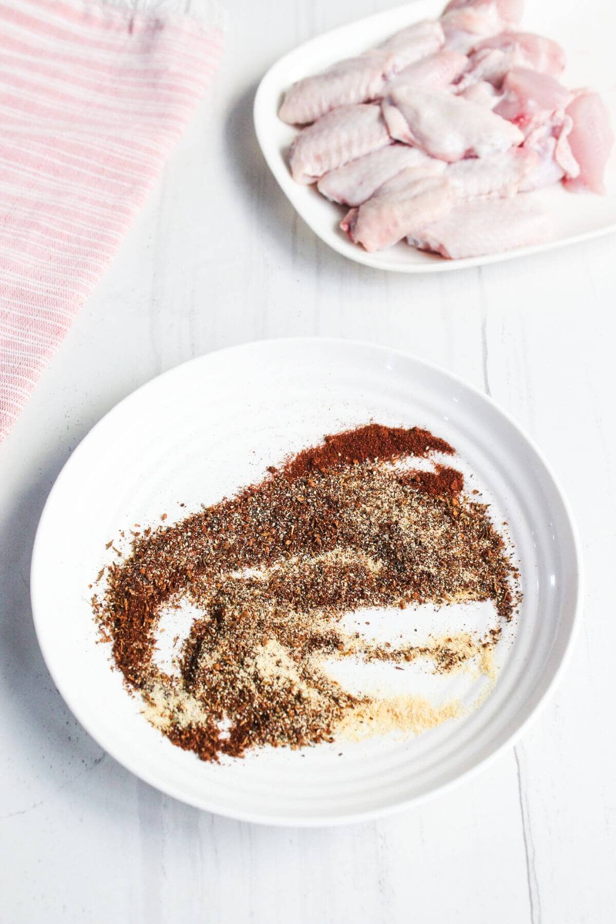 A white plate with assorted ground spices is in the foreground, and a plate of raw chicken wings is in the background on a white surface.