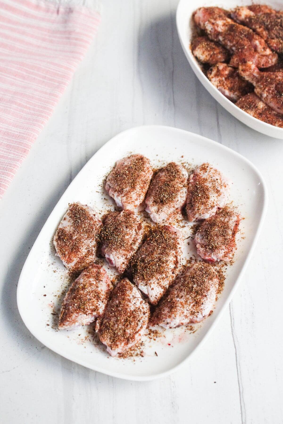Raw chicken wings coated with dry spice seasoning are arranged on a white plate, with a bowl of additional seasoned wings in the background. A pink-striped towel is on the side.