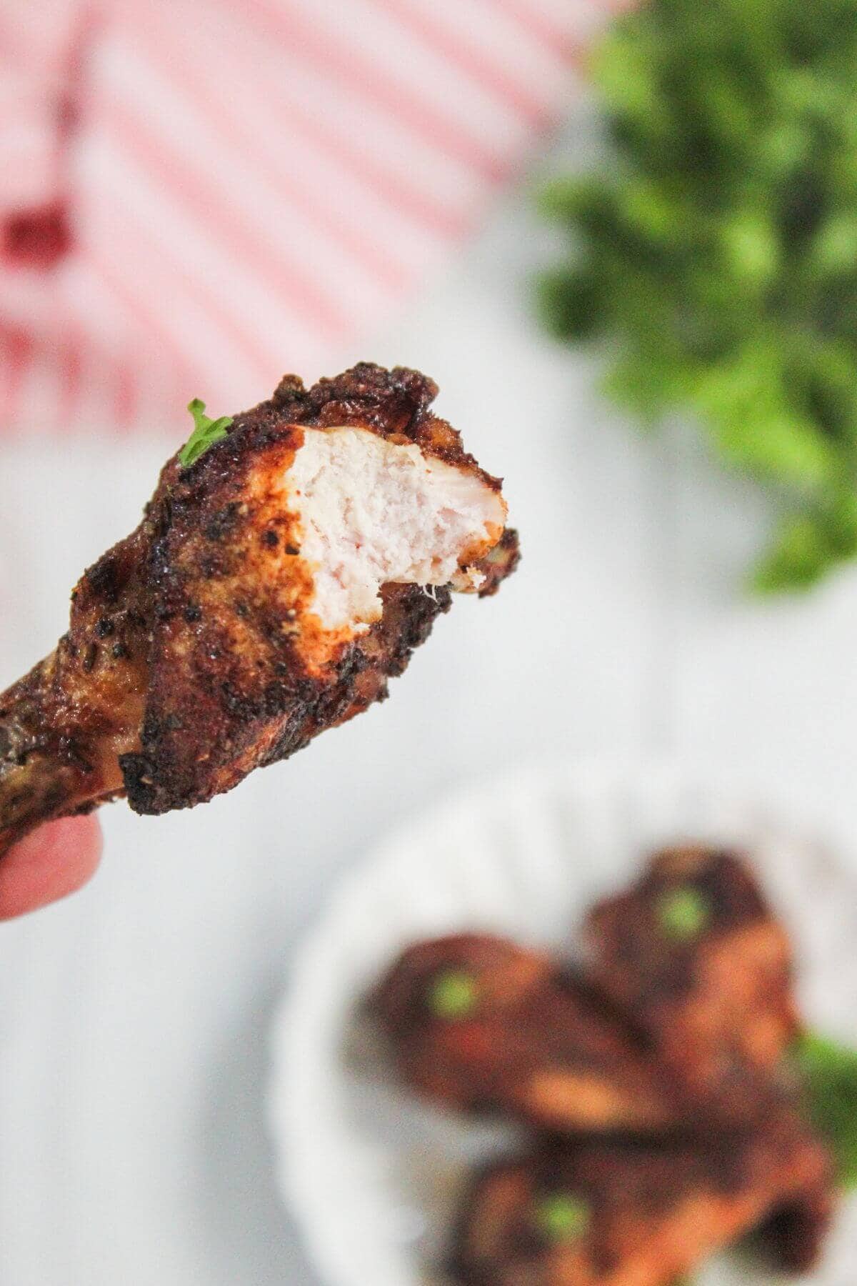 A close-up of a partially eaten, seasoned chicken drumstick being held, with more drumsticks and greenery blurred in the background.