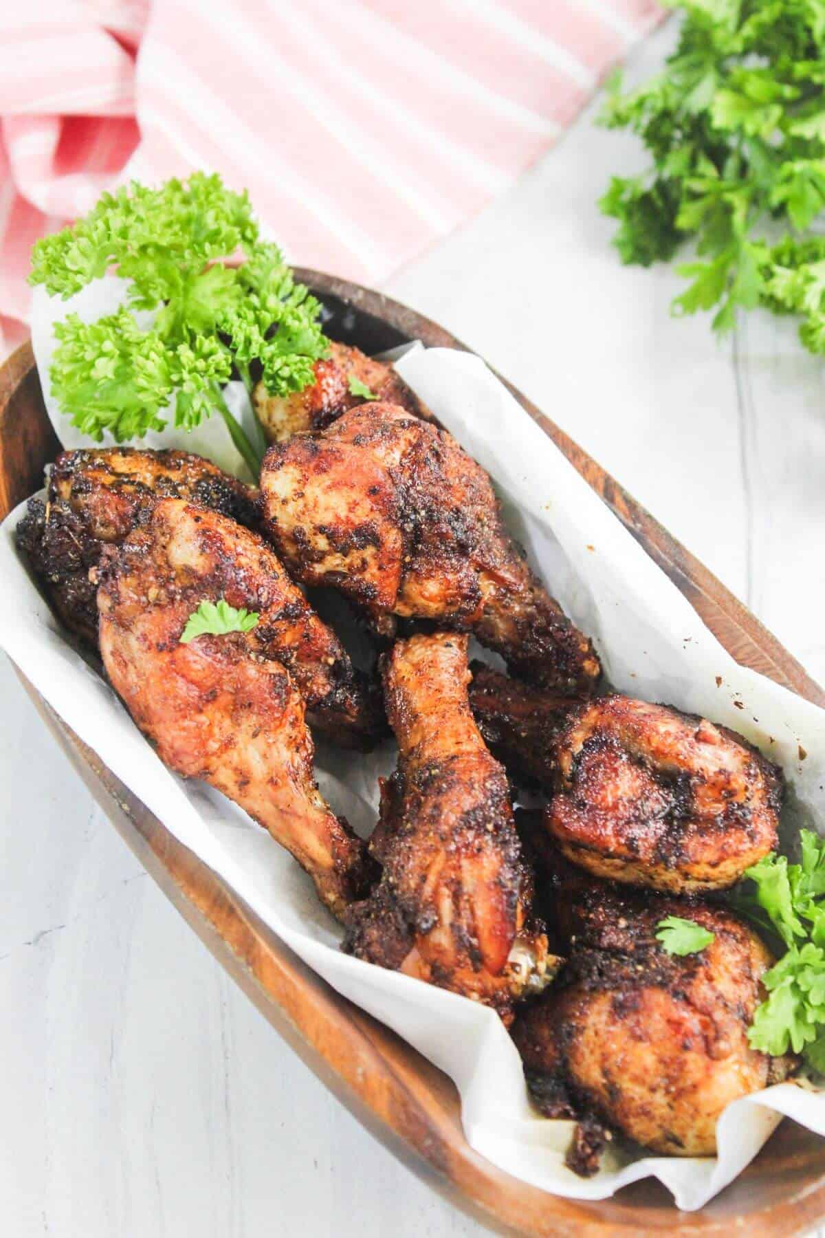 Six pieces of seasoned, cooked chicken drumsticks and wings are arranged in a wooden tray lined with parchment paper, garnished with fresh parsley.