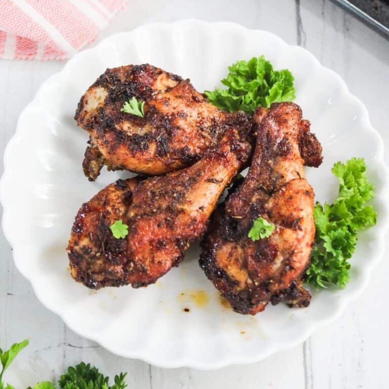 Three seasoned, roasted chicken drumsticks are served on a white scalloped plate, garnished with fresh parsley and small leafy greens.