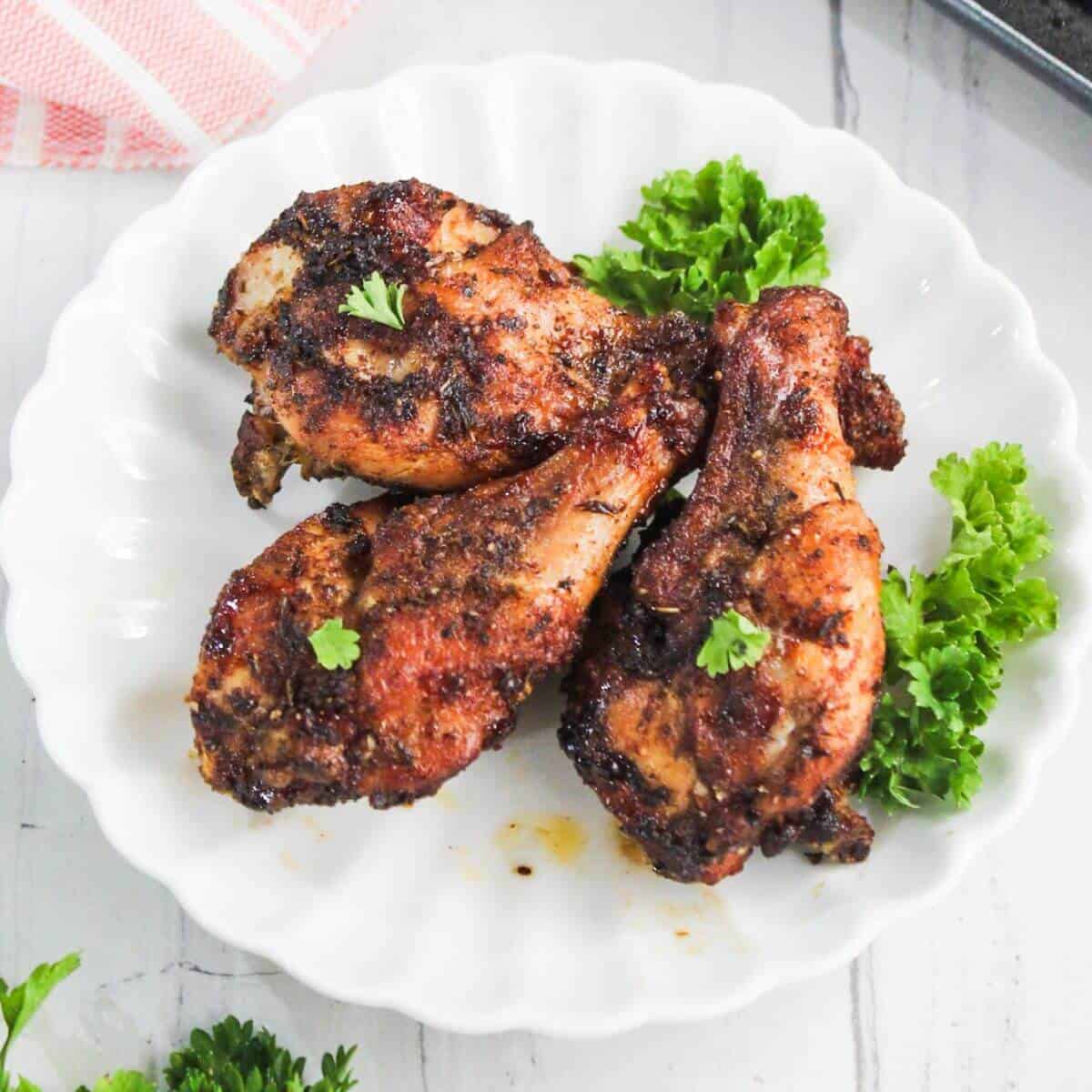 Three seasoned, roasted chicken drumsticks are served on a white scalloped plate, garnished with fresh parsley and small leafy greens.