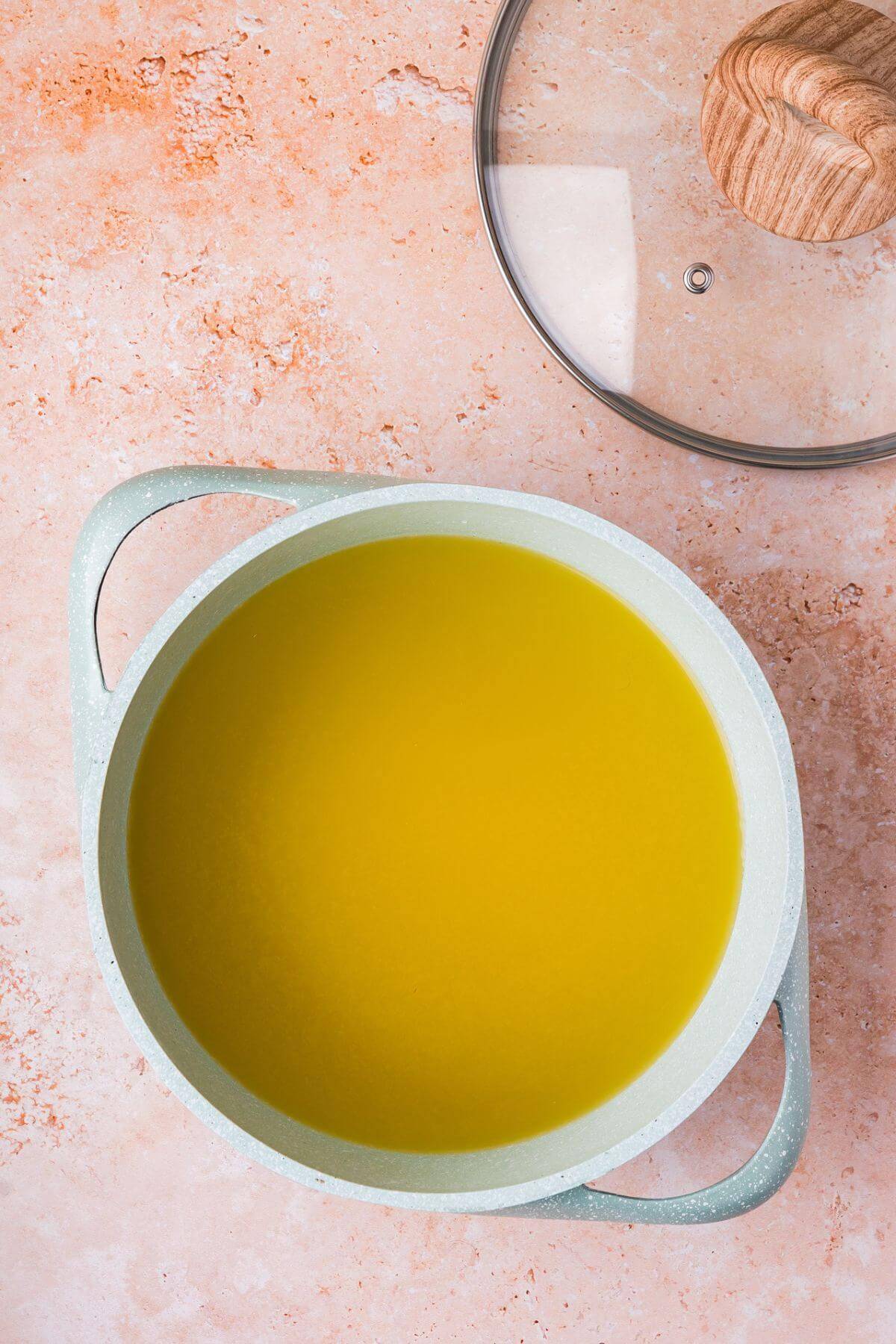 A light green pot filled with yellow liquid sits on a light-colored countertop next to a lid with a wooden handle.