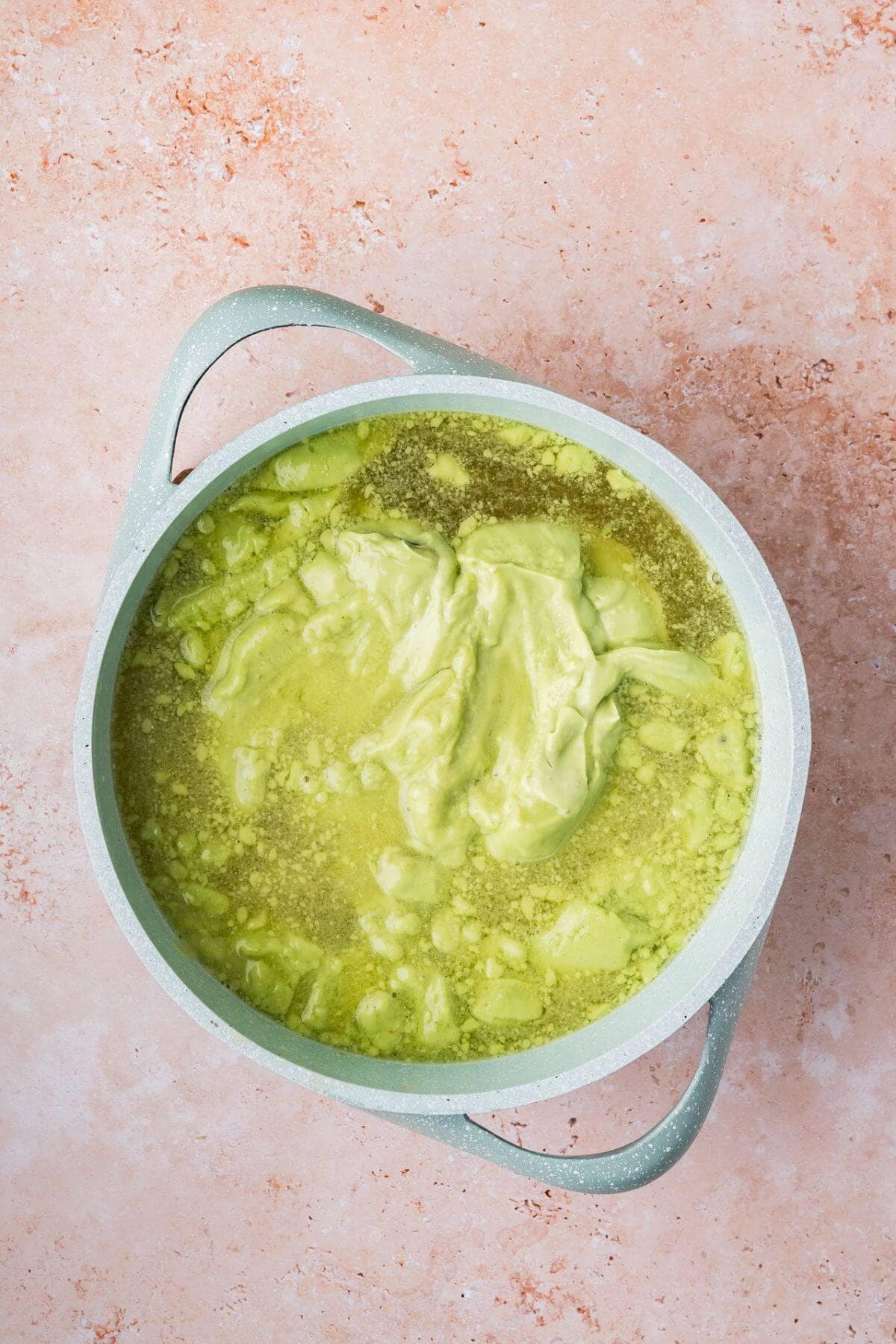 A light green pot filled with a mixture of green and yellow liquid and thick green paste, set on a pink textured surface.