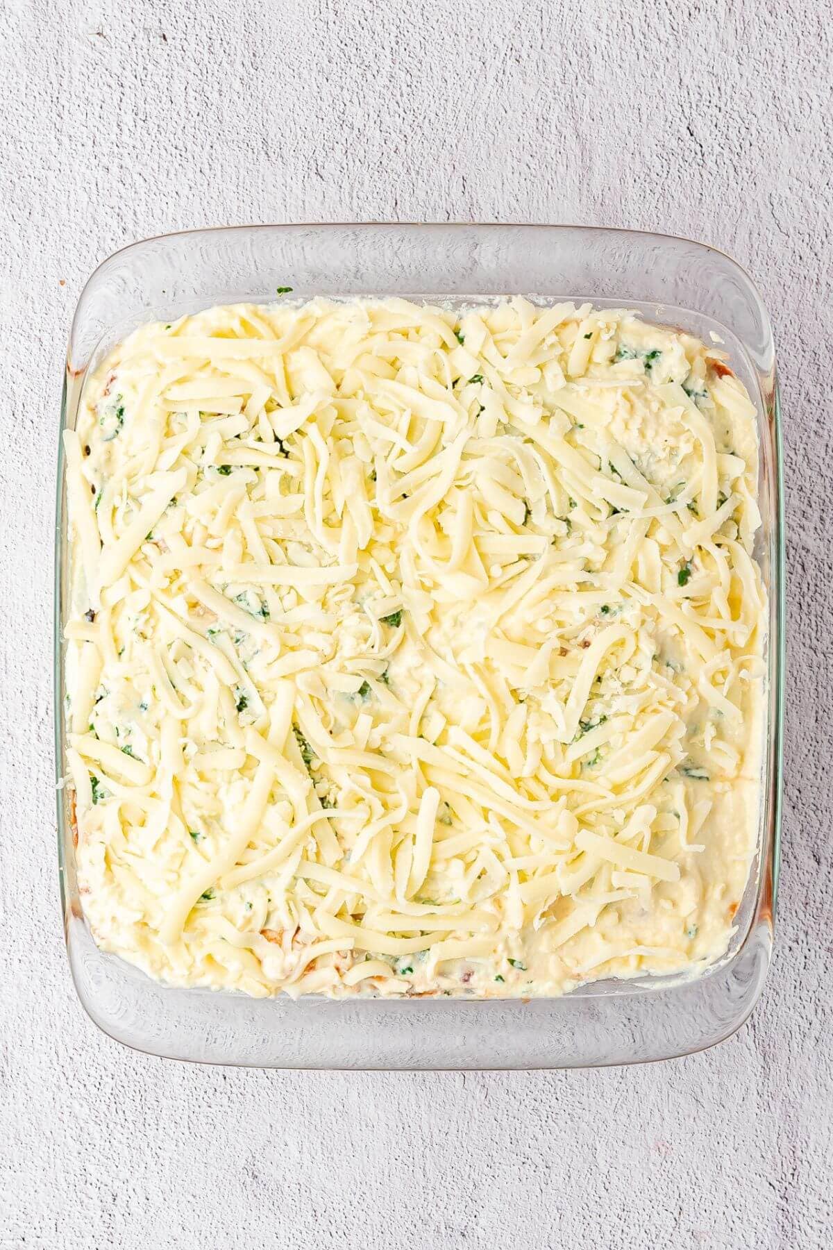 A glass baking dish filled with an unbaked casserole topped with shredded cheese on a light textured surface.
