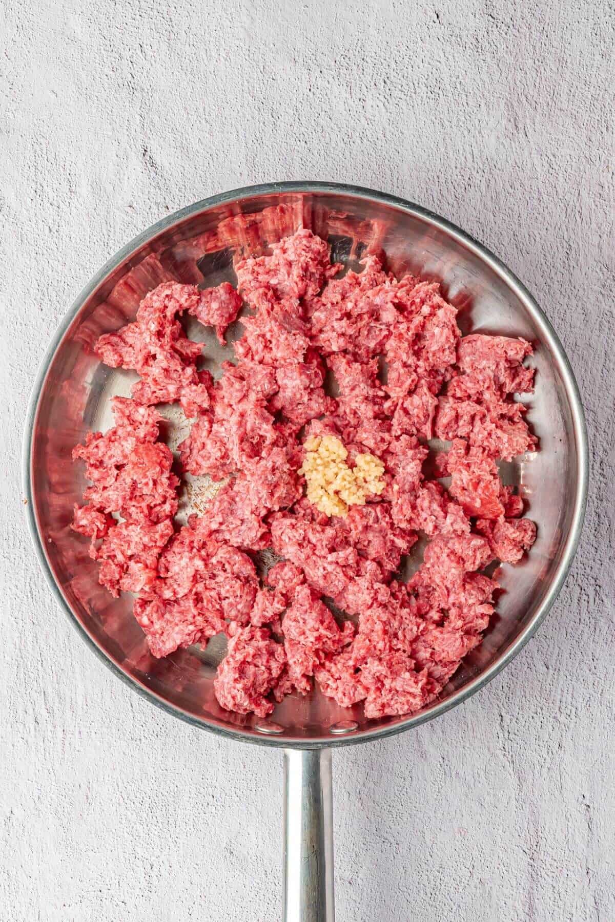 Ground beef and minced garlic in a metal frying pan on a light gray surface, shown before cooking.