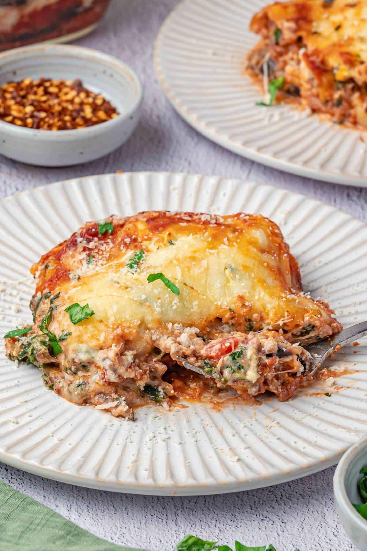 A serving of lasagna with melted cheese and sauce sits on a white plate, partially eaten, with a fork and garnished with chopped herbs.