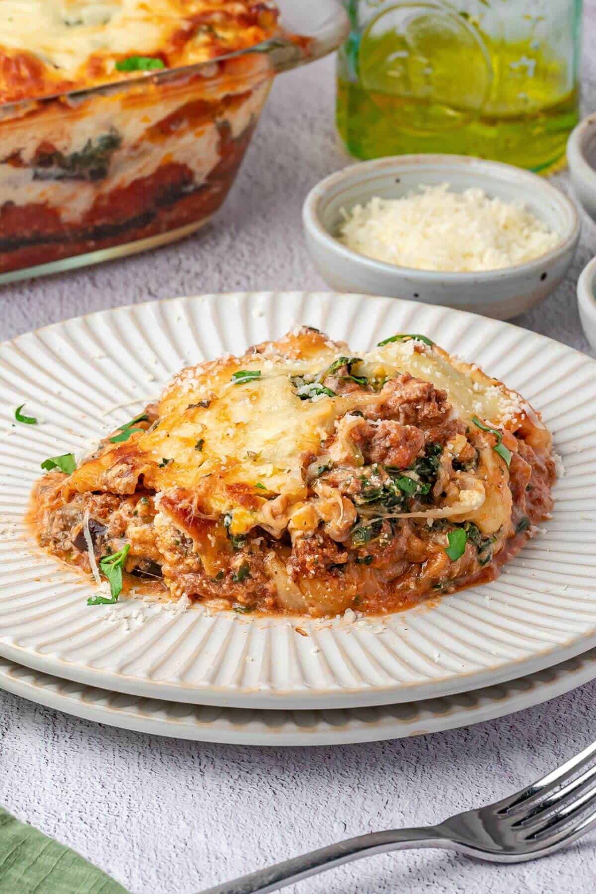 A serving of baked lasagna with cheese, meat, and spinach sits on a white plate, with grated cheese and a baking dish in the background.