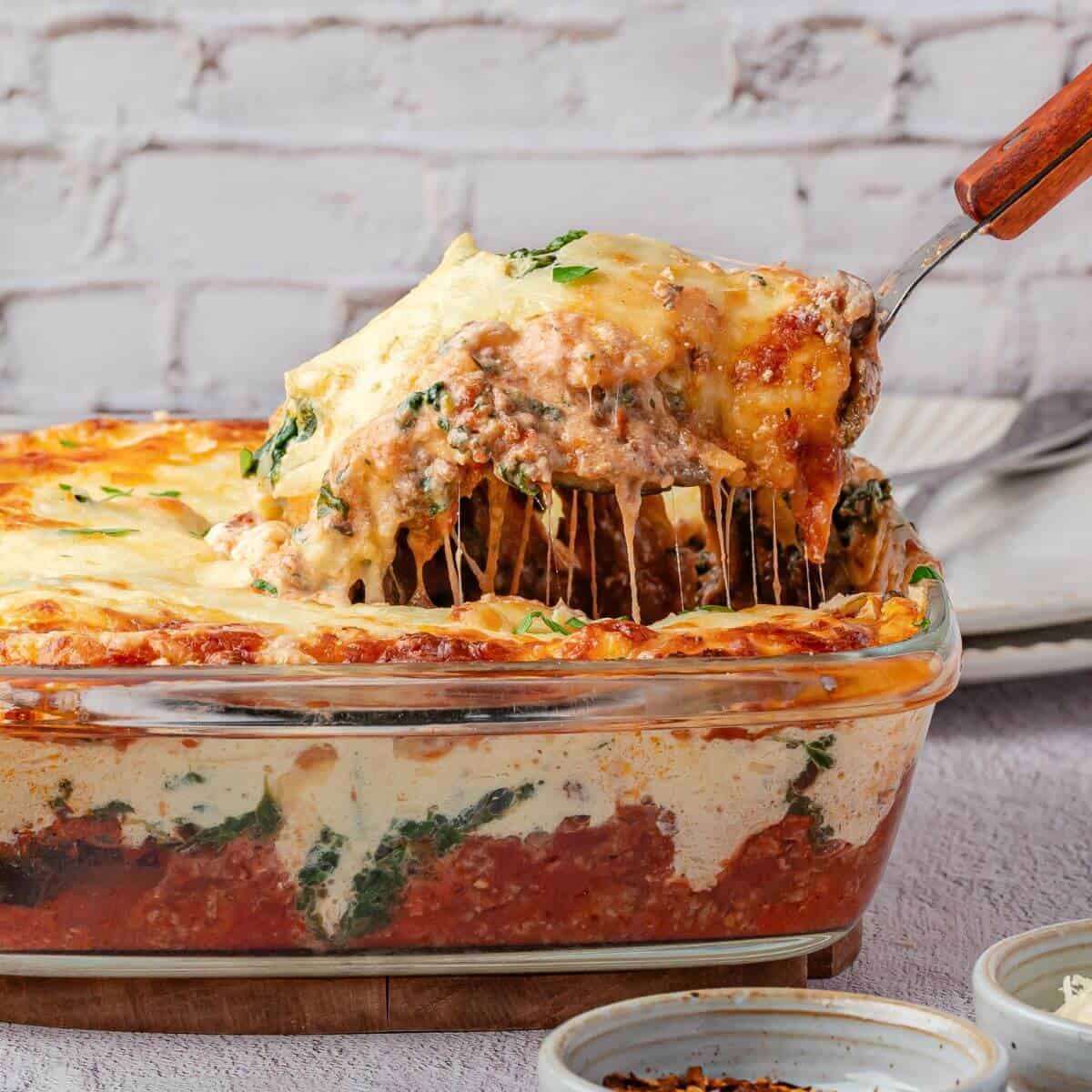 A spatula lifts a cheesy slice of baked lasagna from a glass dish, revealing layers of pasta, sauce, cheese, and spinach.