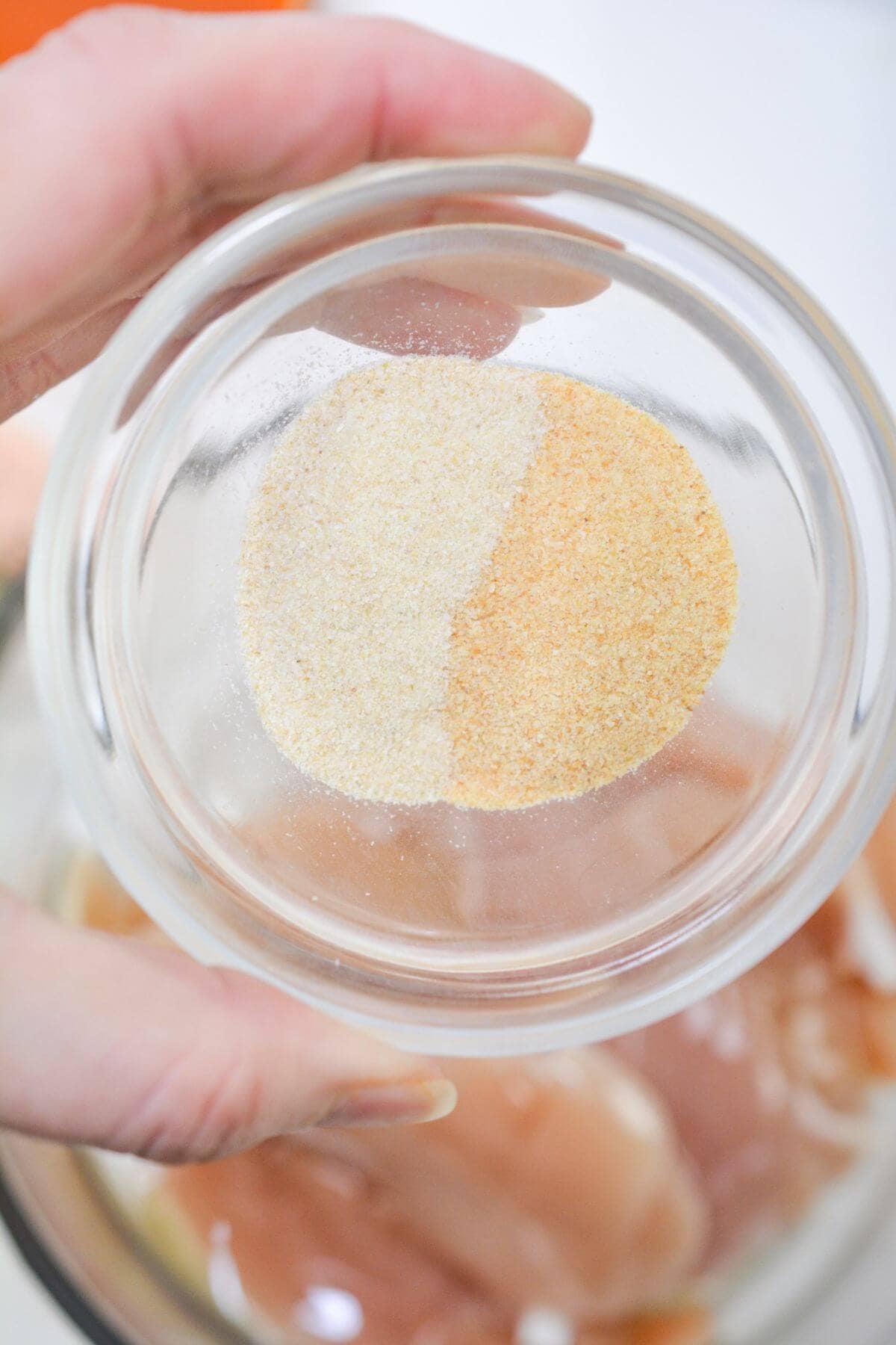 Hand holding a small round glass bowl containing two types of powdered seasoning, one white and one orange, above a bowl of raw chicken.