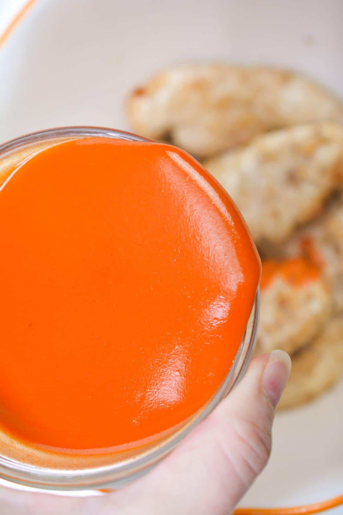 A hand holding a bowl of bright orange sauce above cooked chicken pieces on a white plate.