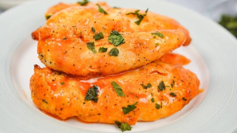 Four seasoned and sauced chicken breast fillets garnished with chopped herbs on a white plate.