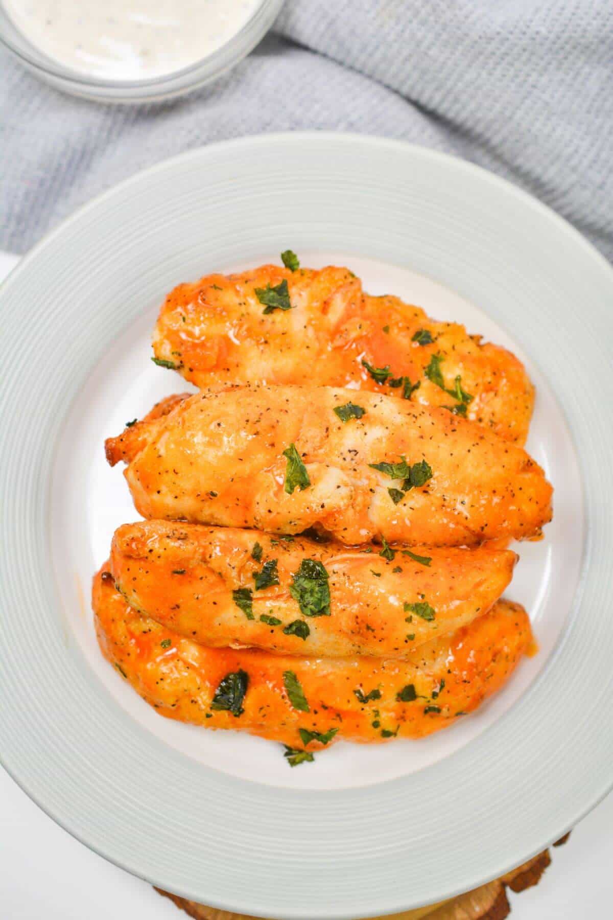 Four seasoned chicken tenders garnished with herbs on a white plate, with a sauce dish in the background.