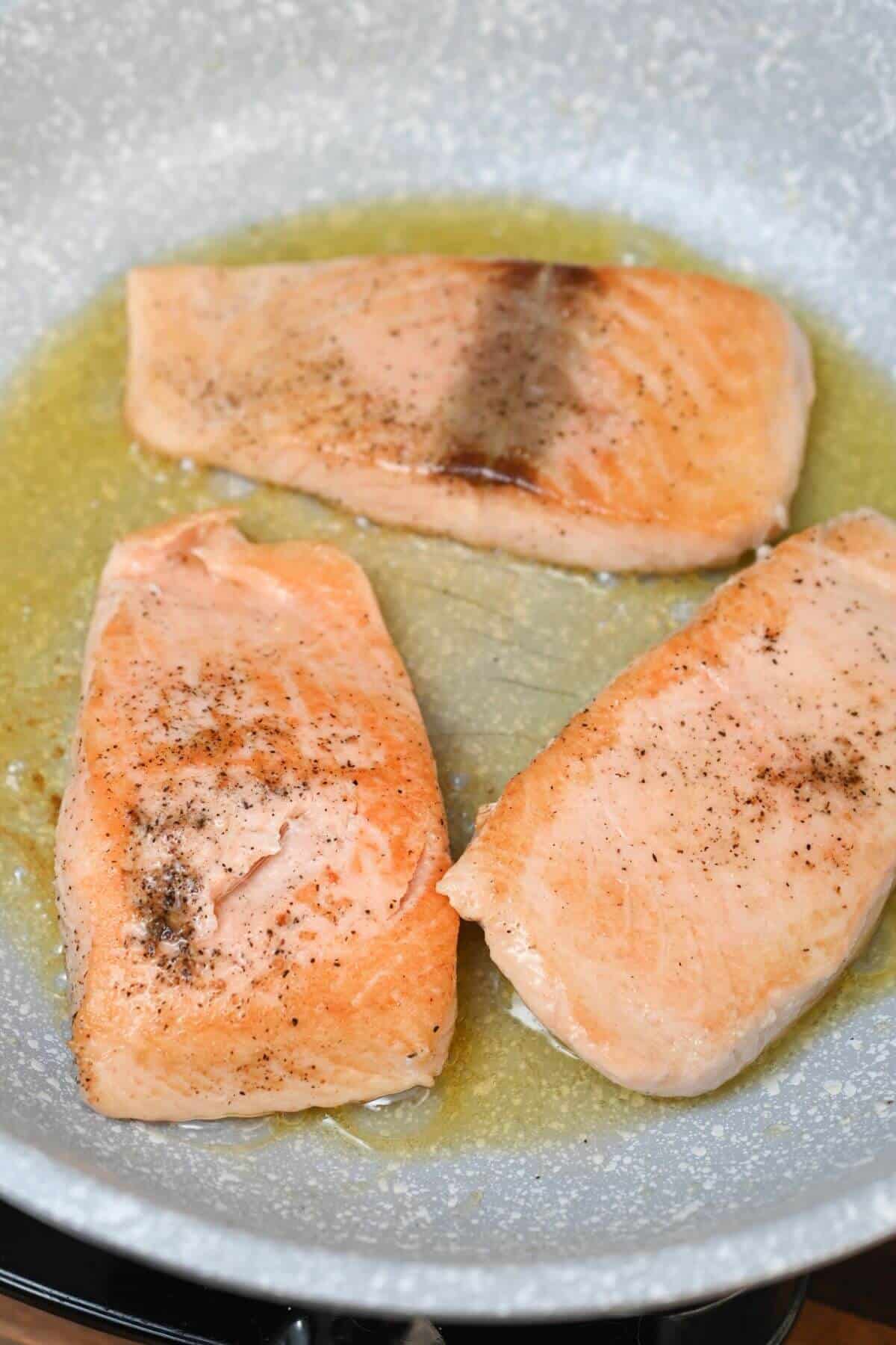 Three salmon fillets cooking in a frying pan with oil, seasoned with pepper.