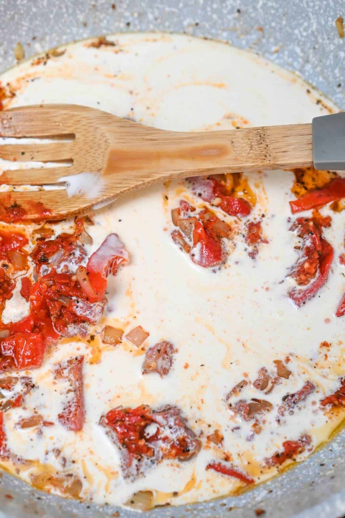 Creamy sauce with cooked tomatoes and spices, sprinkled with herbs, being stirred by a wooden spatula in a pan.