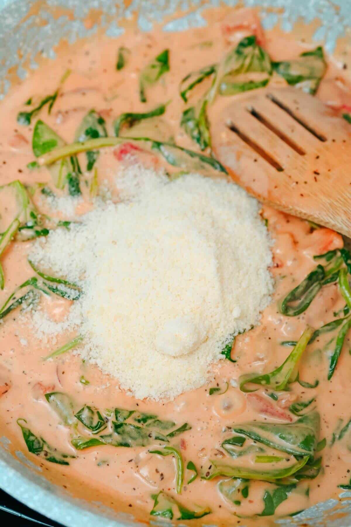 Creamy sauce with spinach, tomatoes, and a sprinkle of parmesan cheese being stirred with a wooden spoon in a pan.