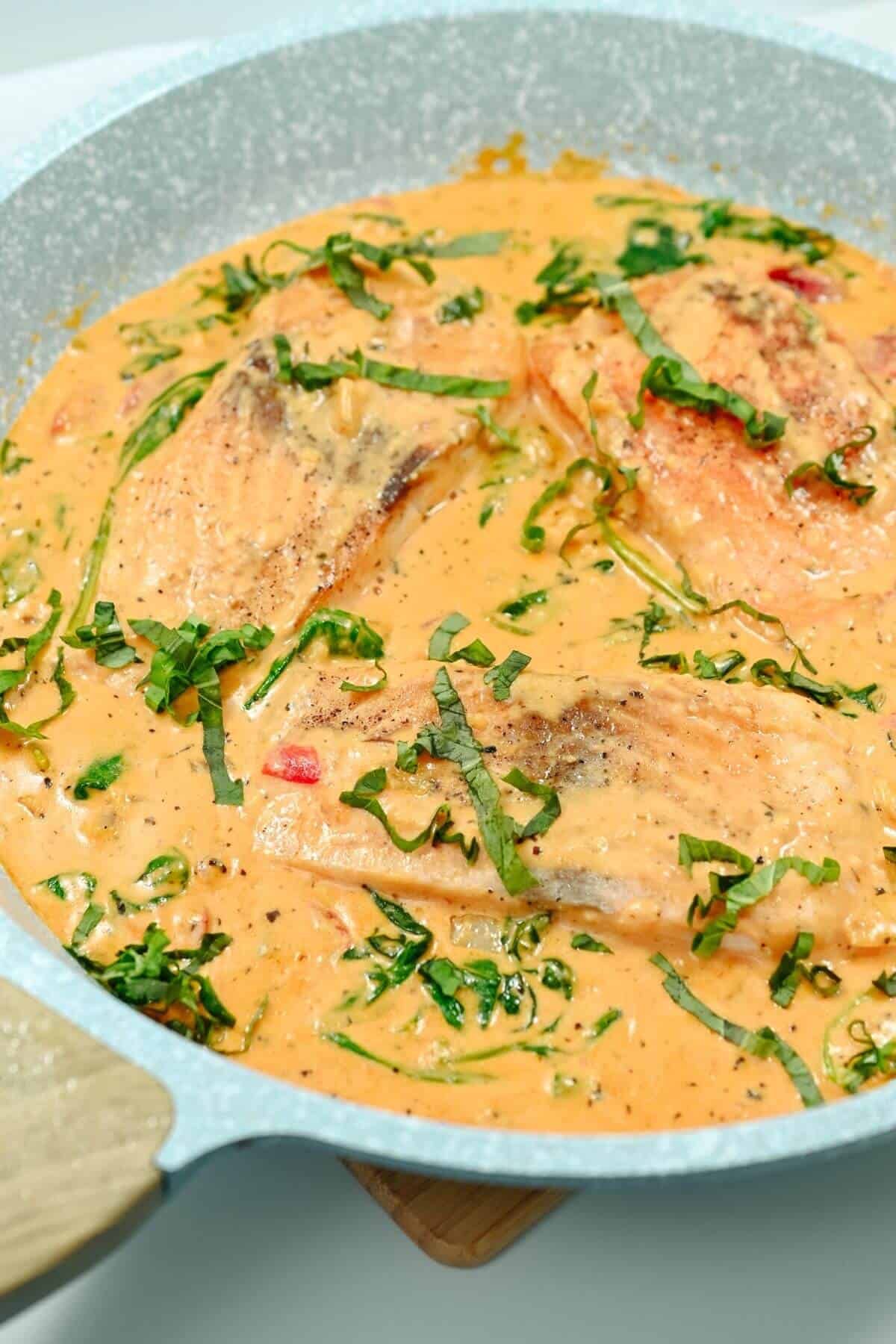 Salmon fillets in a creamy, orange-colored sauce with chopped greens and herbs in a skillet.