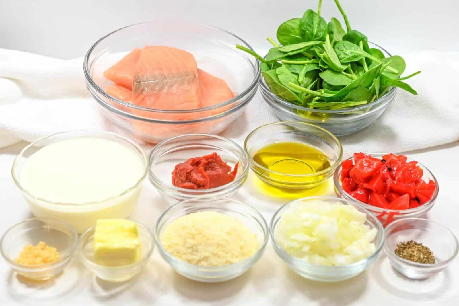 Bowls of raw salmon, spinach, cream, tomato paste, red peppers, olive oil, garlic, butter, Parmesan, onion, and spices on a white surface.