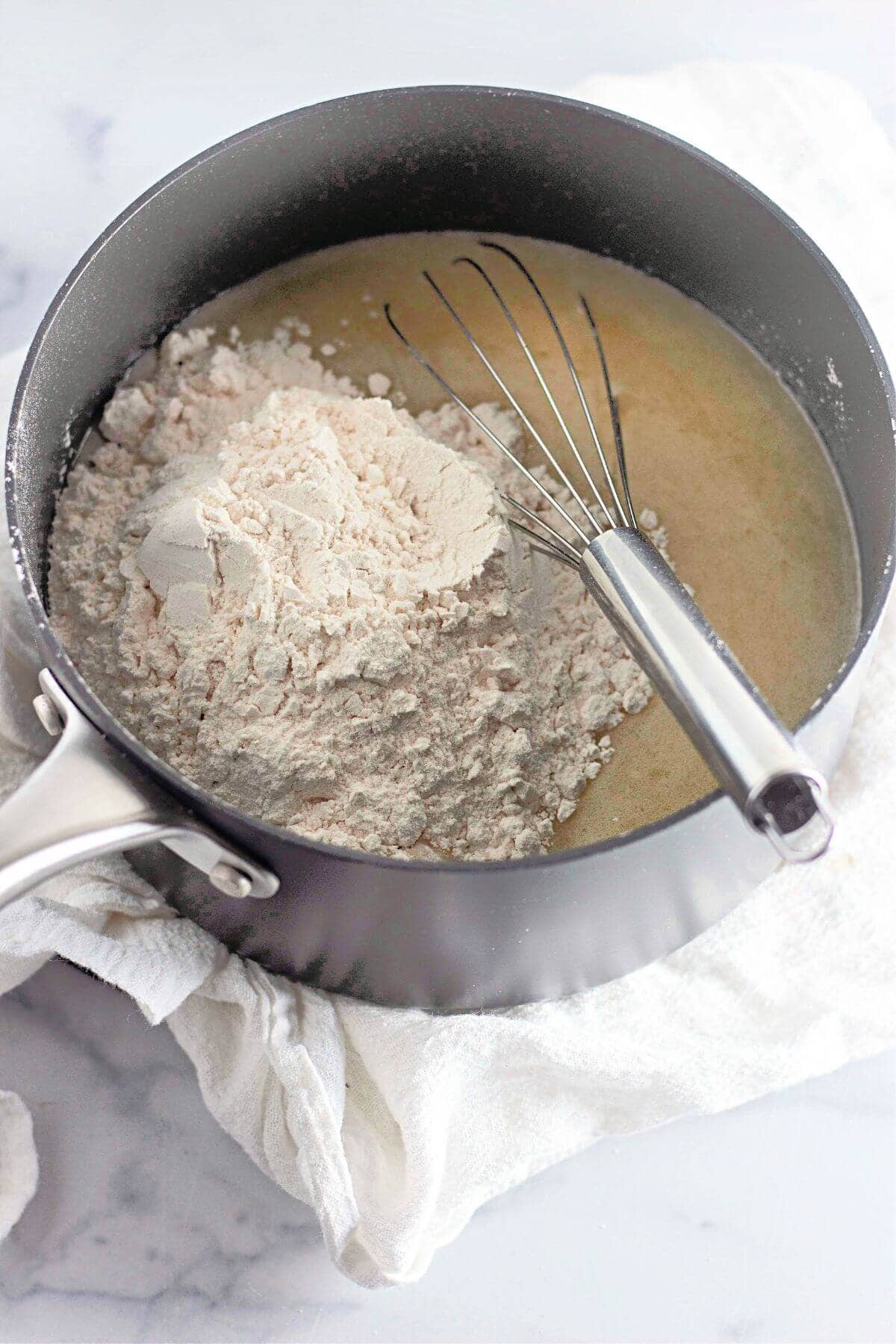 A saucepan with flour and a sauce mixture, placed on a white cloth. A metal whisk rests inside.