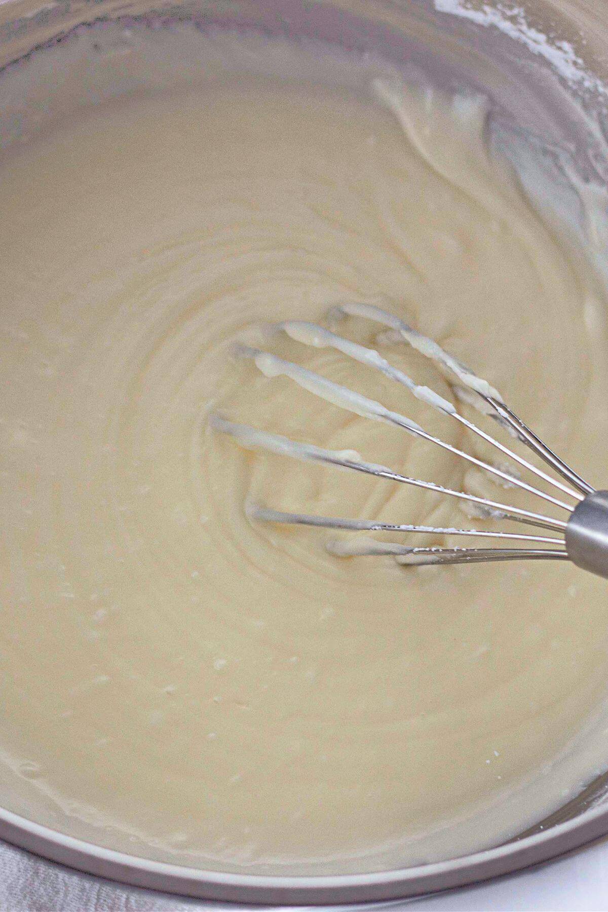 A whisk stirring a smooth, light-colored batter in a mixing bowl.