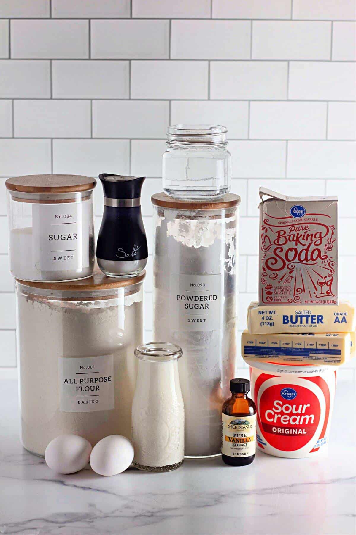 Ingredients for baking: flour, sugar, powdered sugar, baking soda, butter, sour cream, eggs, milk, vanilla extract, and salt on a marble surface with a white tile background.