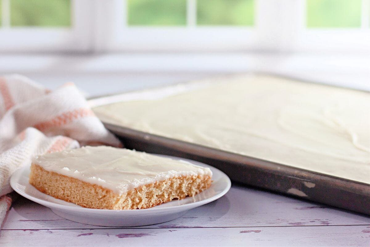 A slice of frosted sheet cake on a plate rests beside a baking tray with more cake. A striped cloth is next to the plate.