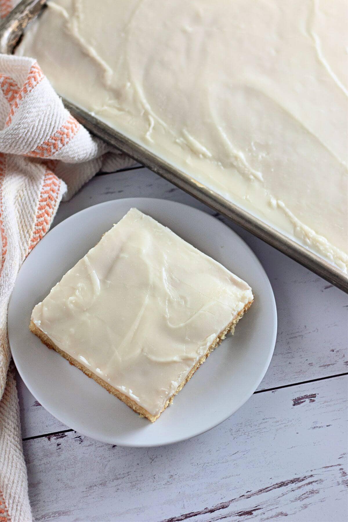 A frosted sheet cake with a cut square piece on a white plate, next to an orange and white patterned cloth.