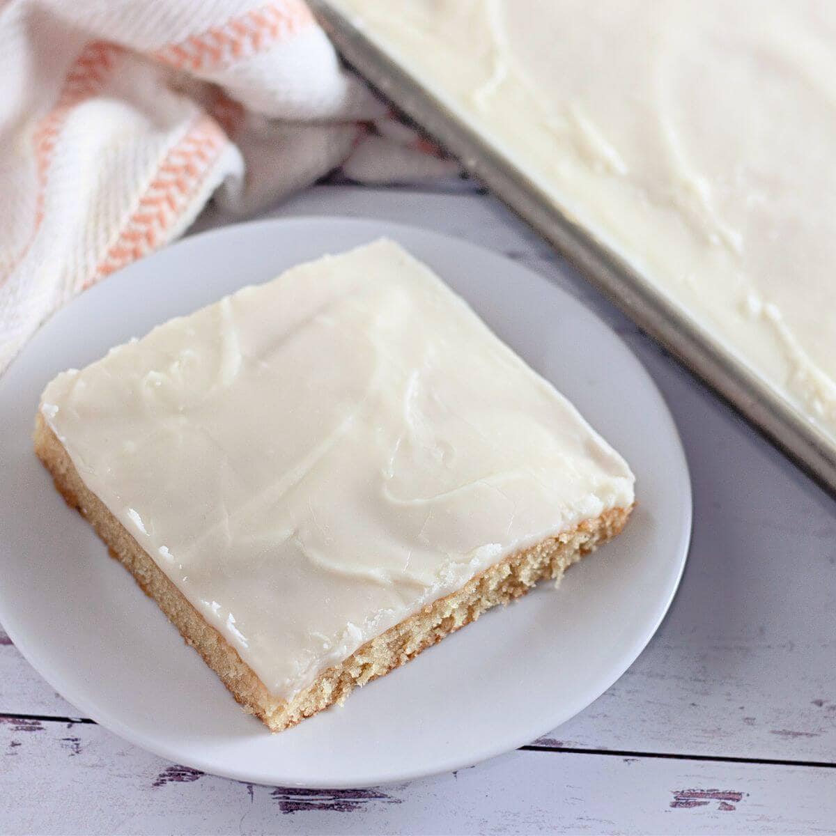 A square piece of white frosted cake on a white plate, with more cake in a pan and a cloth napkin nearby.