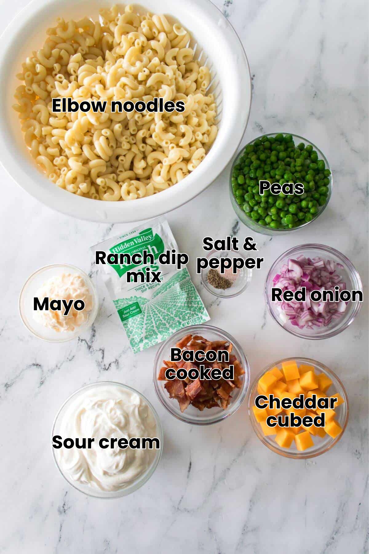 Ingredients for a pasta salad, including cooked macaroni, peas, red onion, cheddar cheese, bacon, ranch dip mix, mayonnaise, and sour cream, arranged in bowls on a marble surface.