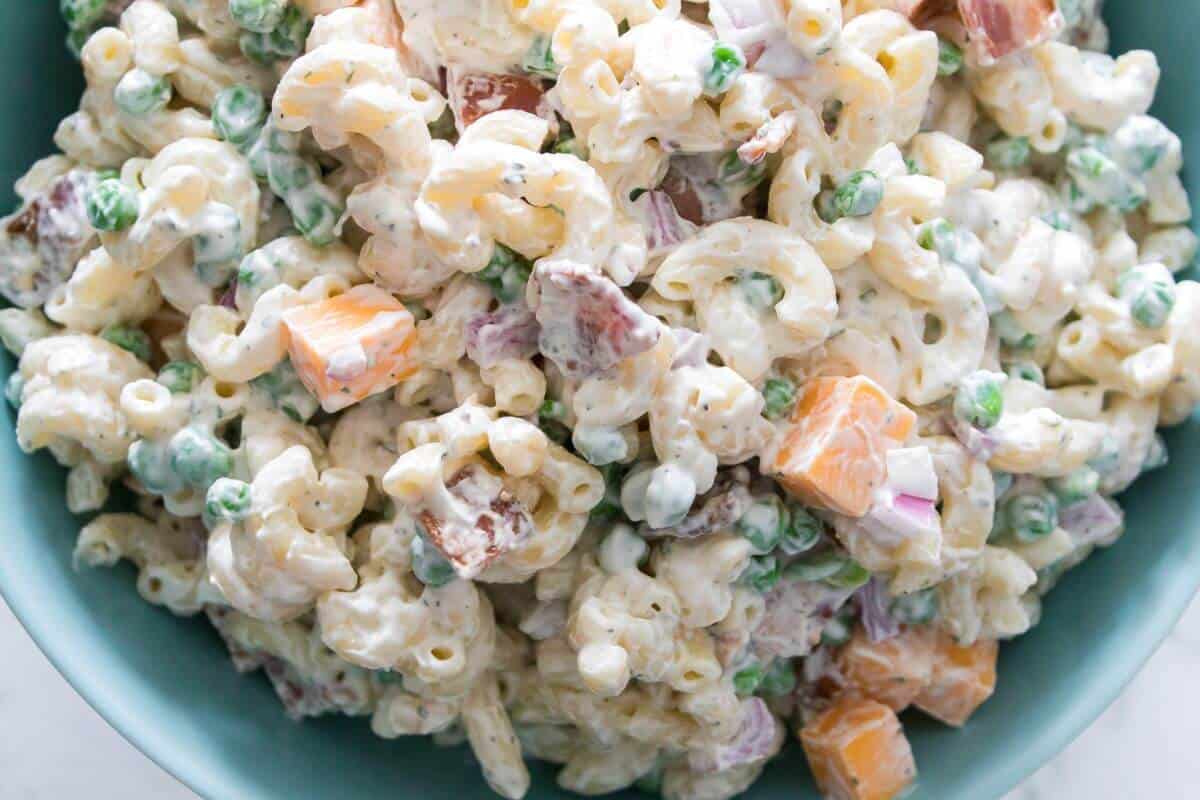 A close-up of macaroni salad with peas, cubed cheddar cheese, diced red onion, and bacon pieces, all mixed in a creamy dressing.