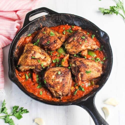 A cast iron skillet with five seasoned chicken thighs in tomato sauce, garnished with herbs, sits on a white surface near fresh parsley, garlic cloves, and pepper mills.