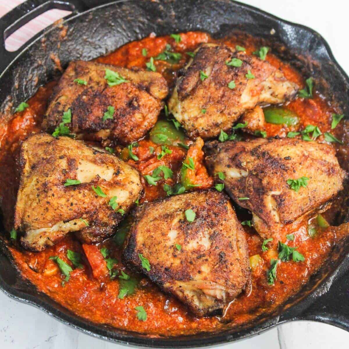 A cast iron skillet filled with seasoned, roasted chicken thighs on top of a chunky tomato and bell pepper sauce, garnished with chopped herbs.