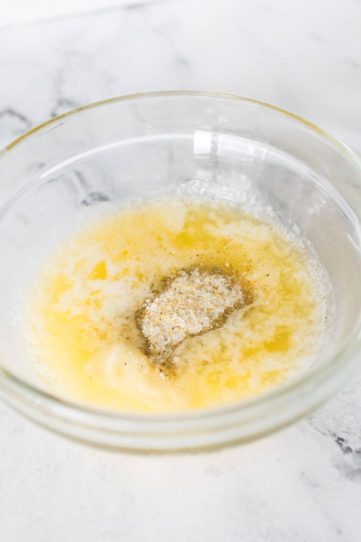 A clear glass bowl containing melted butter mixed with seasonings and breadcrumbs on a white marble surface.