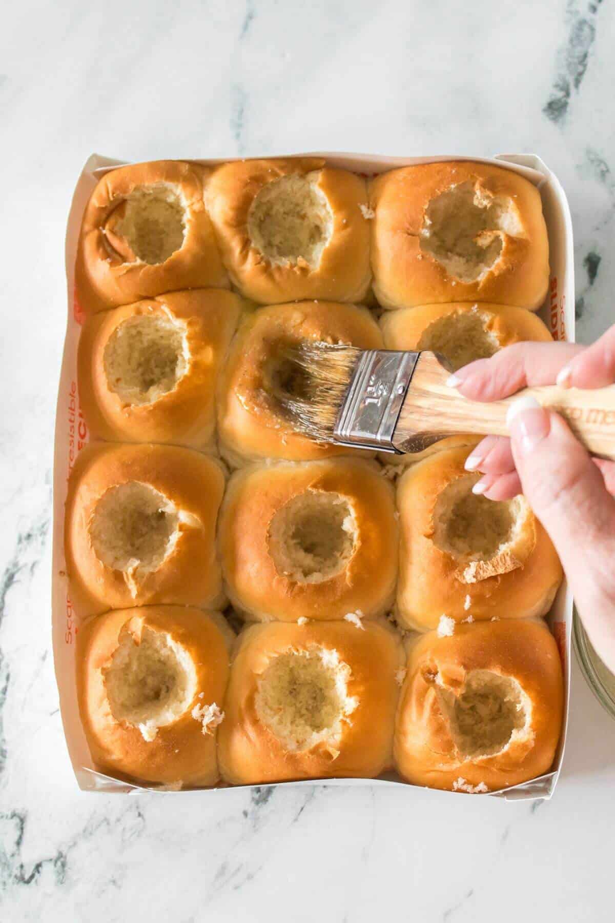 A hand uses a brush to apply liquid to the tops of twelve bread rolls, each with a hollowed-out center, arranged in a baking tray on a marble surface.