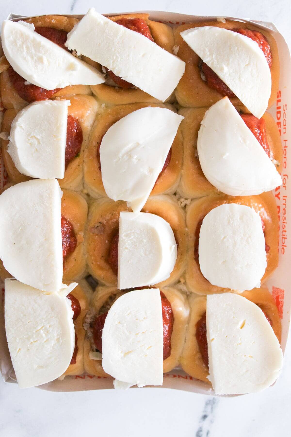 A tray of slider buns topped with tomato sauce and thick slices of mozzarella cheese, arranged in a grid pattern on a white surface.