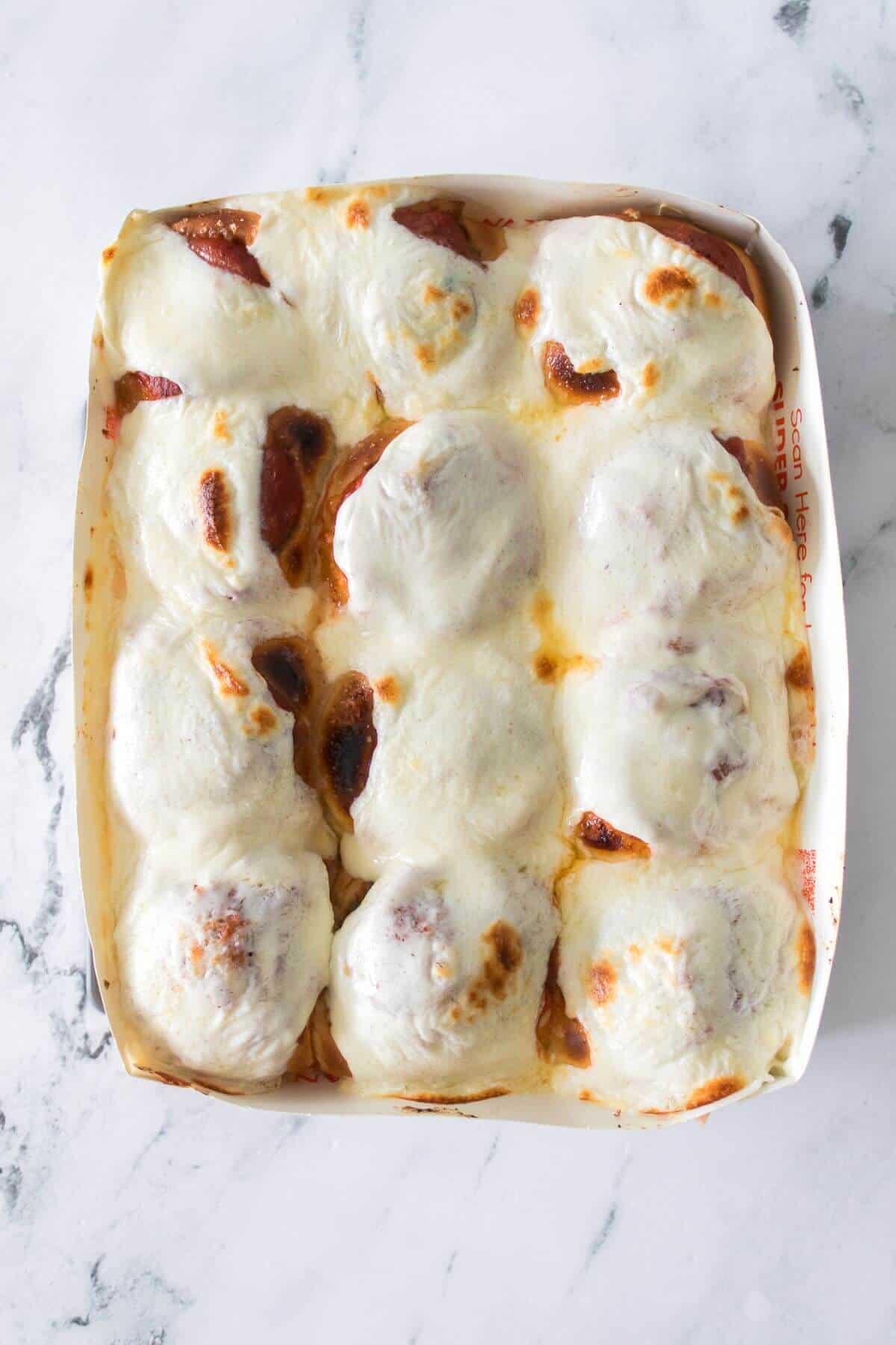 A baking dish containing nine meatballs covered with melted, browned mozzarella cheese on a marble surface.