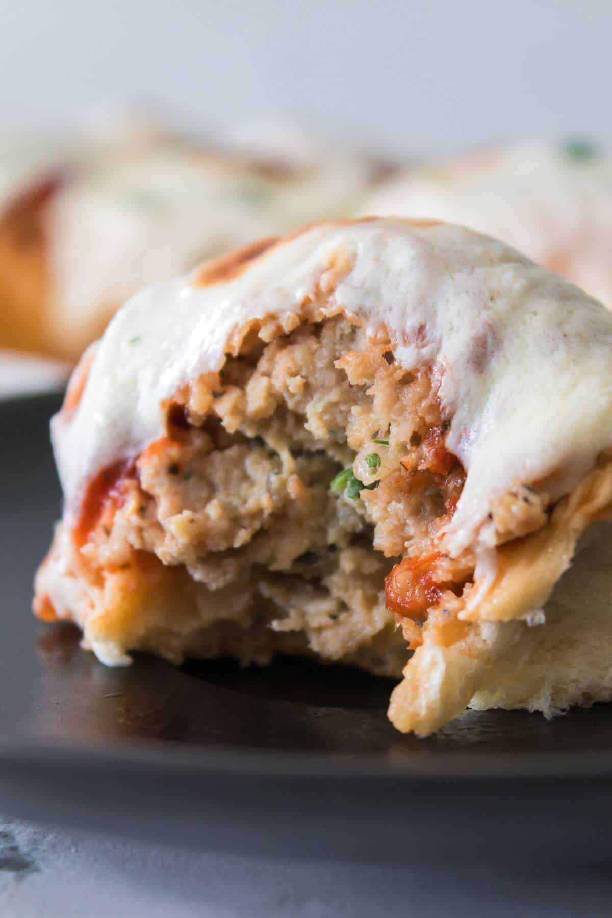 A close-up of a baked stuffed bread roll filled with a meat mixture, topped with melted cheese, and shown with a bite taken out.