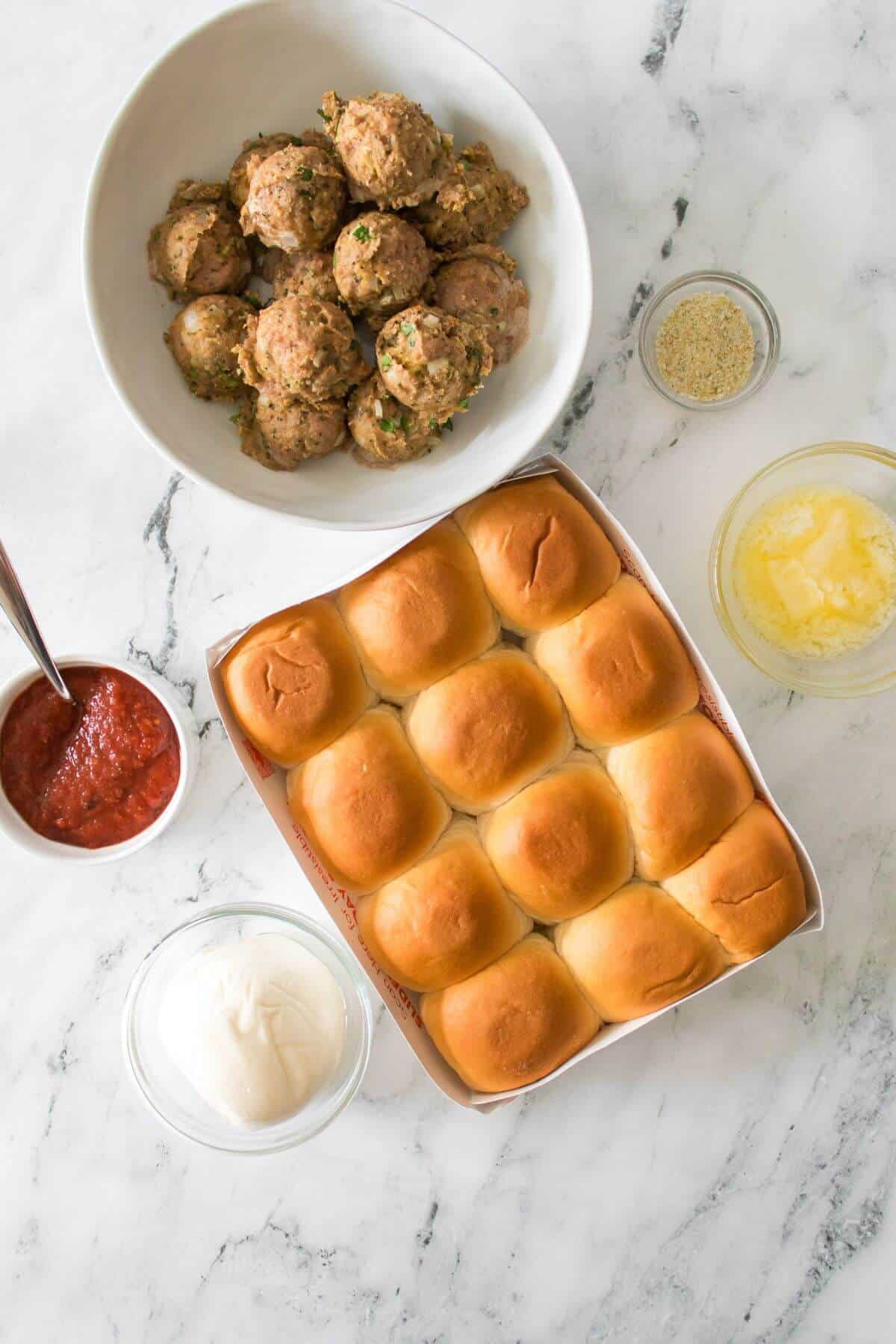A tray of twelve slider buns, a bowl of cooked meatballs, a bowl of marinara sauce, shredded cheese, melted butter, and breadcrumbs on a marble surface.